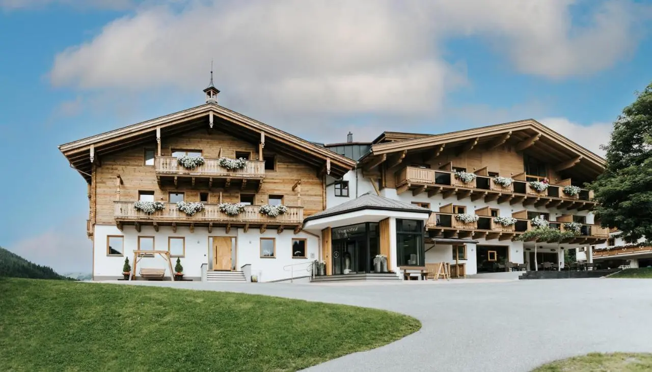 Traditionelles Alpenhotel Thurnerhof mit Holzbalkonen und Blumenkästen vor grüner Wiese und blauem Himmel.