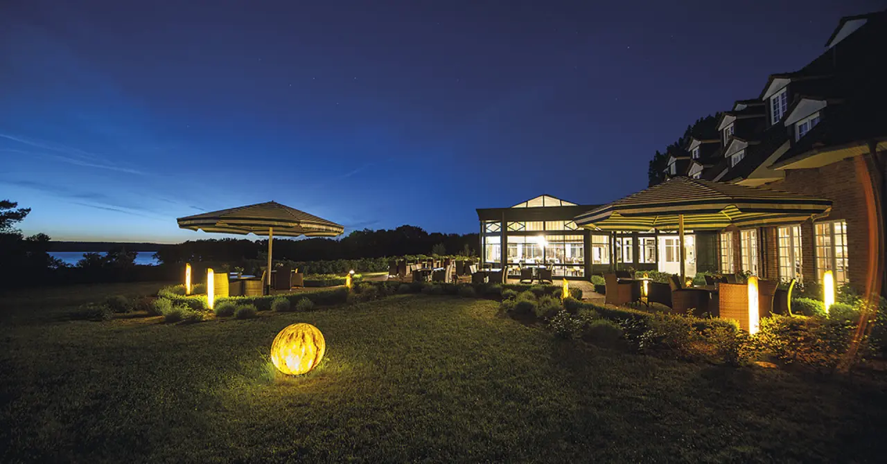 Abendliche Aussenansicht des Hotel Bornmühle mit beleuchteter Terrasse, Garten und See im Hintergrund unter blauem Nachthimmel.