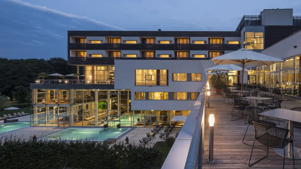 Abendansicht des SPA RESORT STYRIA mit beleuchtetem Aussenpool, Innenbereich und Terrasse unter blauem Himmel.