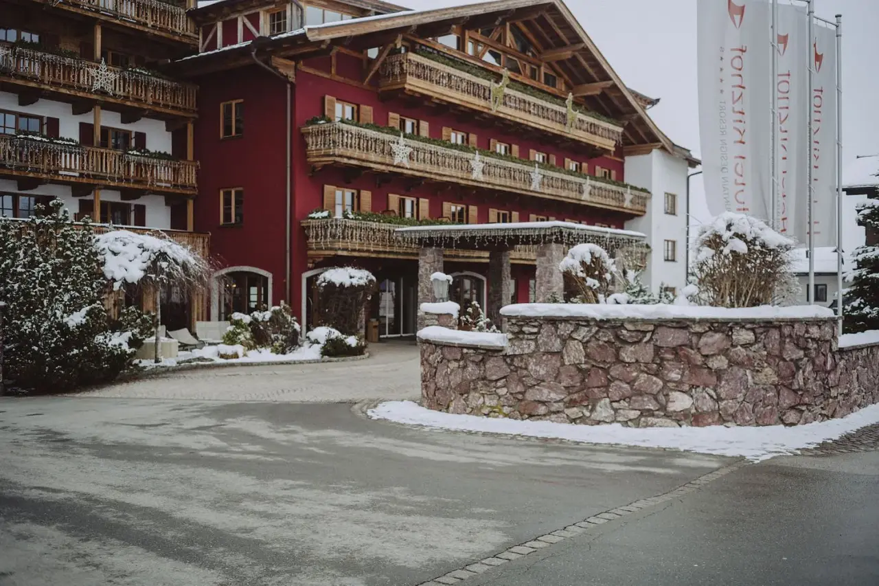 Verschneite Aussenansicht des Hotel Kitzhof Mountain Design Resort in Kitzbühel mit roter Fassade und traditionellen Balkonen.