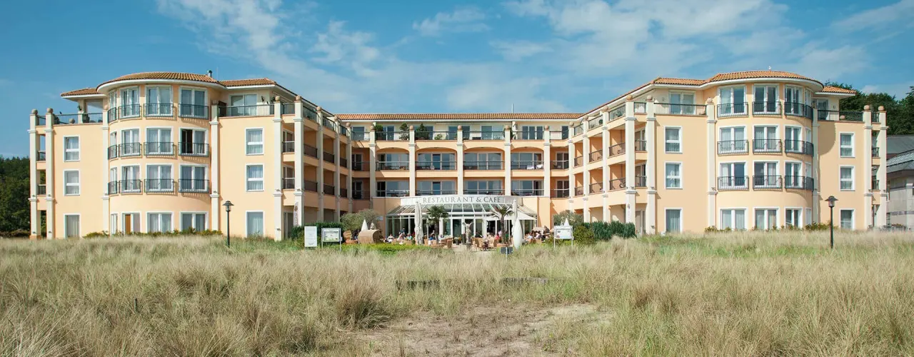 Aussenansicht des Hotel Gran Belveder mit mediterraner Architektur und Restaurantterrasse vor Dünengras und blauem Himmel.