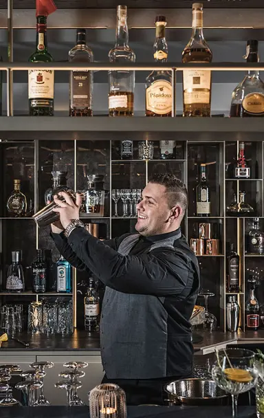 Ein lächelnder Barkeeper mixt einen Cocktail an der gut sortierten Bar des Hotel Post Kaltenbach.