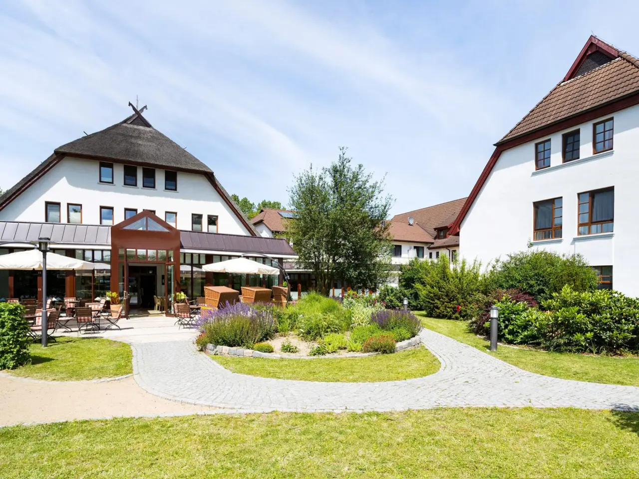 Aussenansicht des Hotel Warnemünder Hof mit traditionellem Reetdach, gepflegtem Garten und einladender Sonnenterrasse.