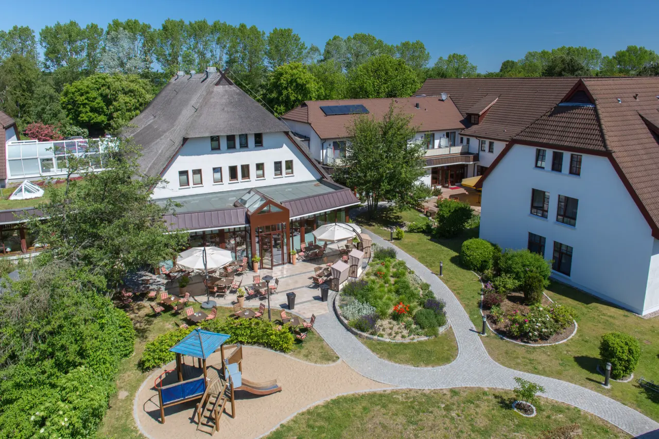 Luftaufnahme des Hotel Warnemünder Hof mit Reetdach, Sonnenterrasse, Garten und Spielplatz unter blauem Himmel.