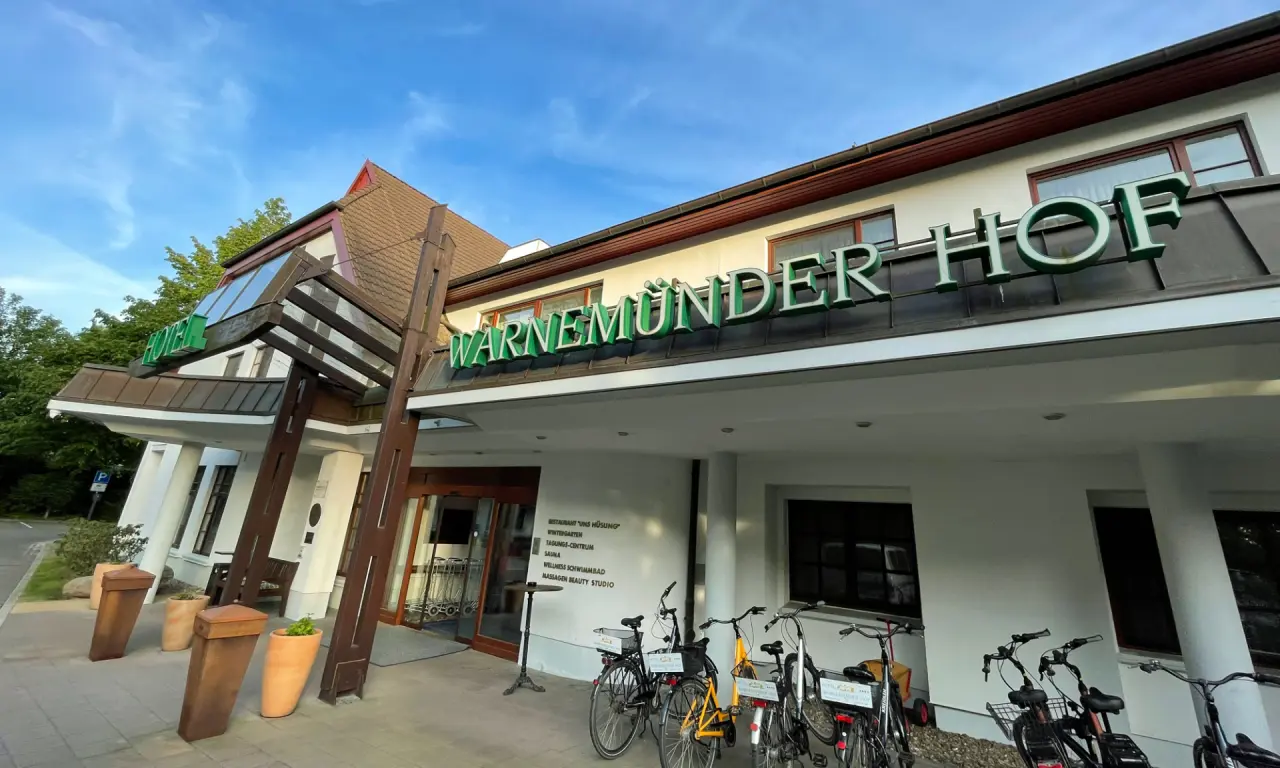 Eingangsbereich des Hotel Warnemünder Hof mit grüner Leuchtschrift und abgestellten Fahrrädern unter blauem Himmel.