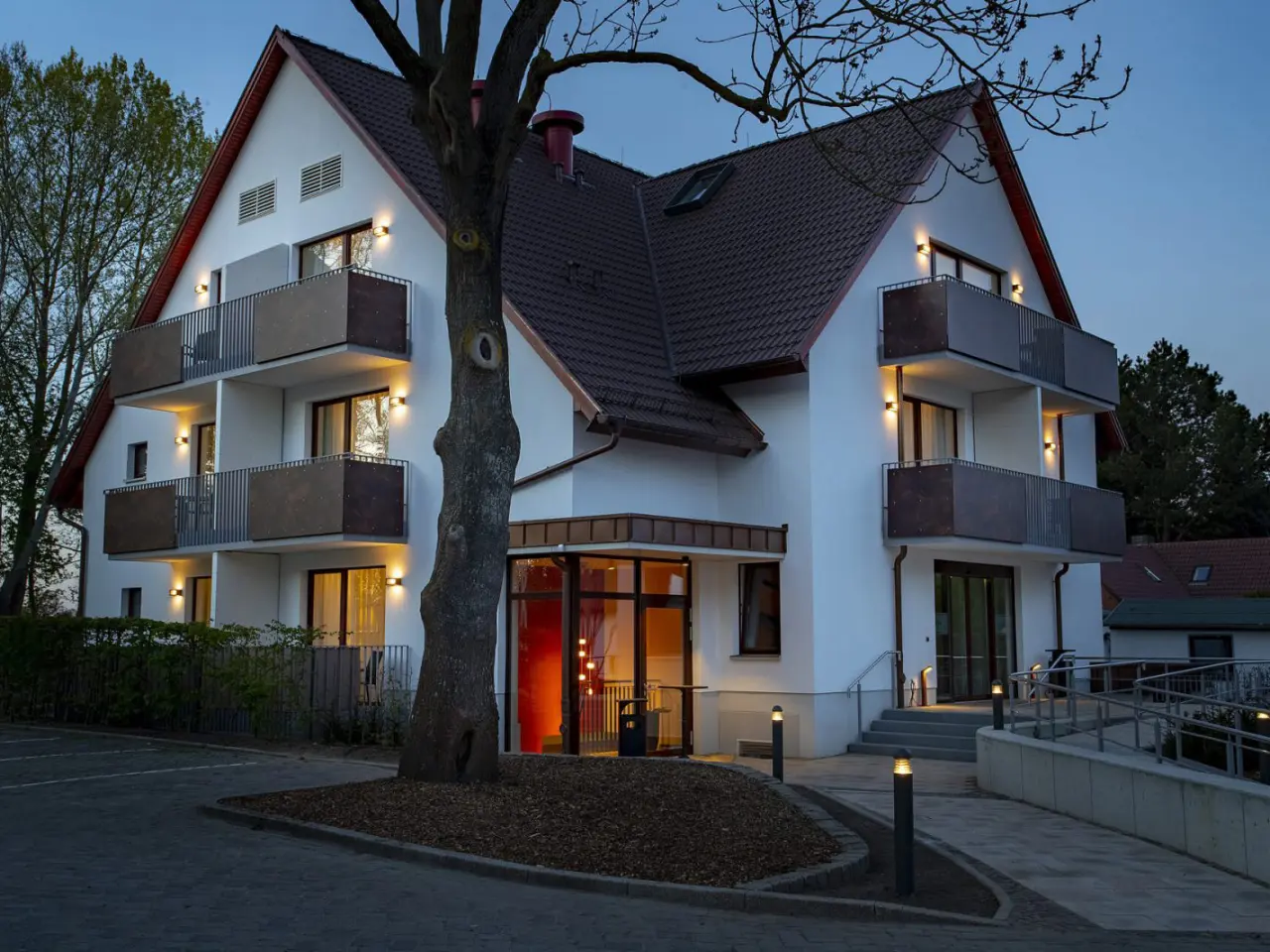 Abendansicht des Hotels Warnemünder Hof mit beleuchteter Fassade und Balkonen, umgeben von Bäumen.