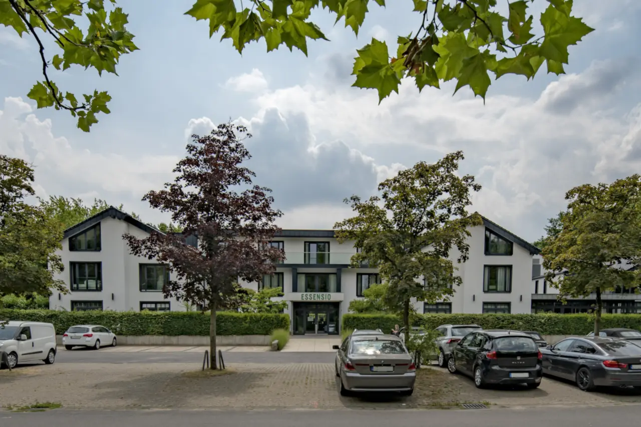 Moderne Aussenansicht des Essensio Hotels mit weisser Fassade, grossen Fenstern und grüner Umgebung unter blauem Himmel.