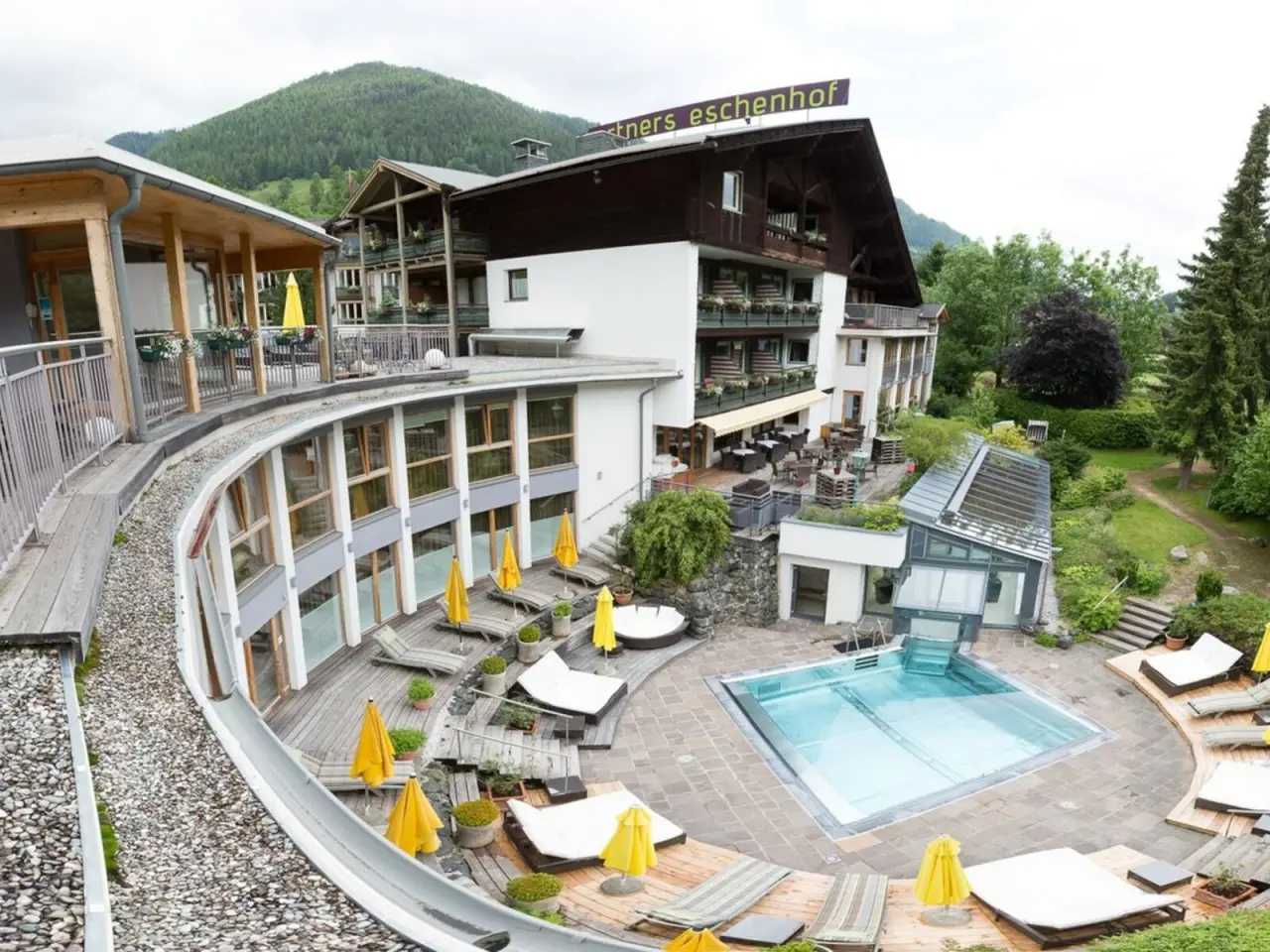 Aussenansicht des Ortners Eschenhof mit geschwungenem Gebäude, Aussenpool und Liegewiese vor bewaldeten Bergen.