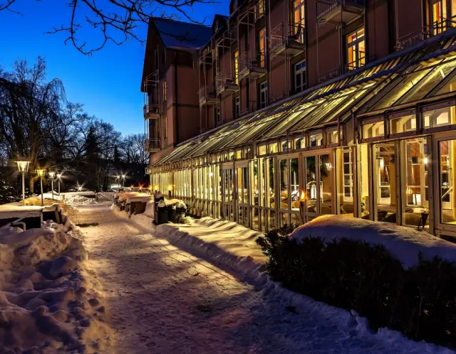 Winterliche Aussenansicht des relexa hotel Bad Steben bei Blaue Stunde. Beleuchteter Eingang und verschneiter Gartenweg.