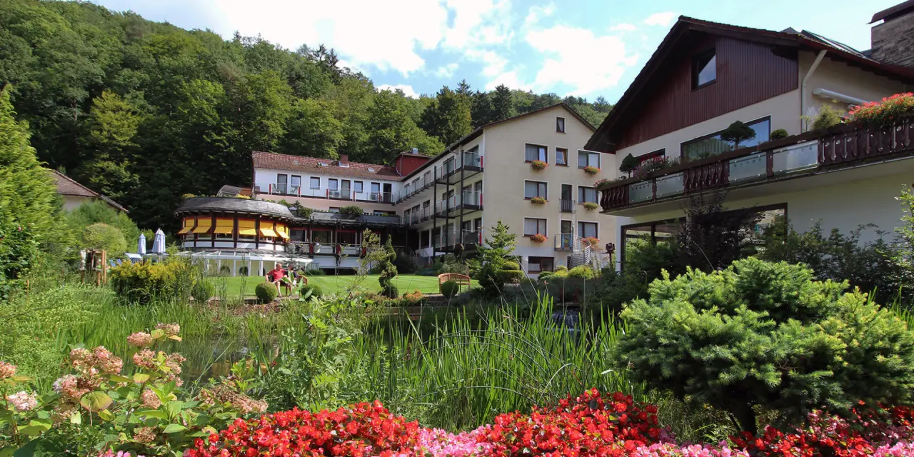 Aussenansicht des Gesundheits- und VitalCenter Gollée mit üppigem Garten, Teich und Wald im Hintergrund.