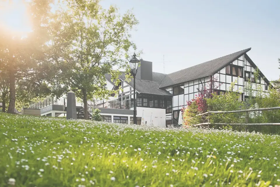 Aussenansicht des Sporthotels Grafenwald mit traditioneller Fachwerkarchitektur und blühender Wiese im Vordergrund bei Sonnenschein.