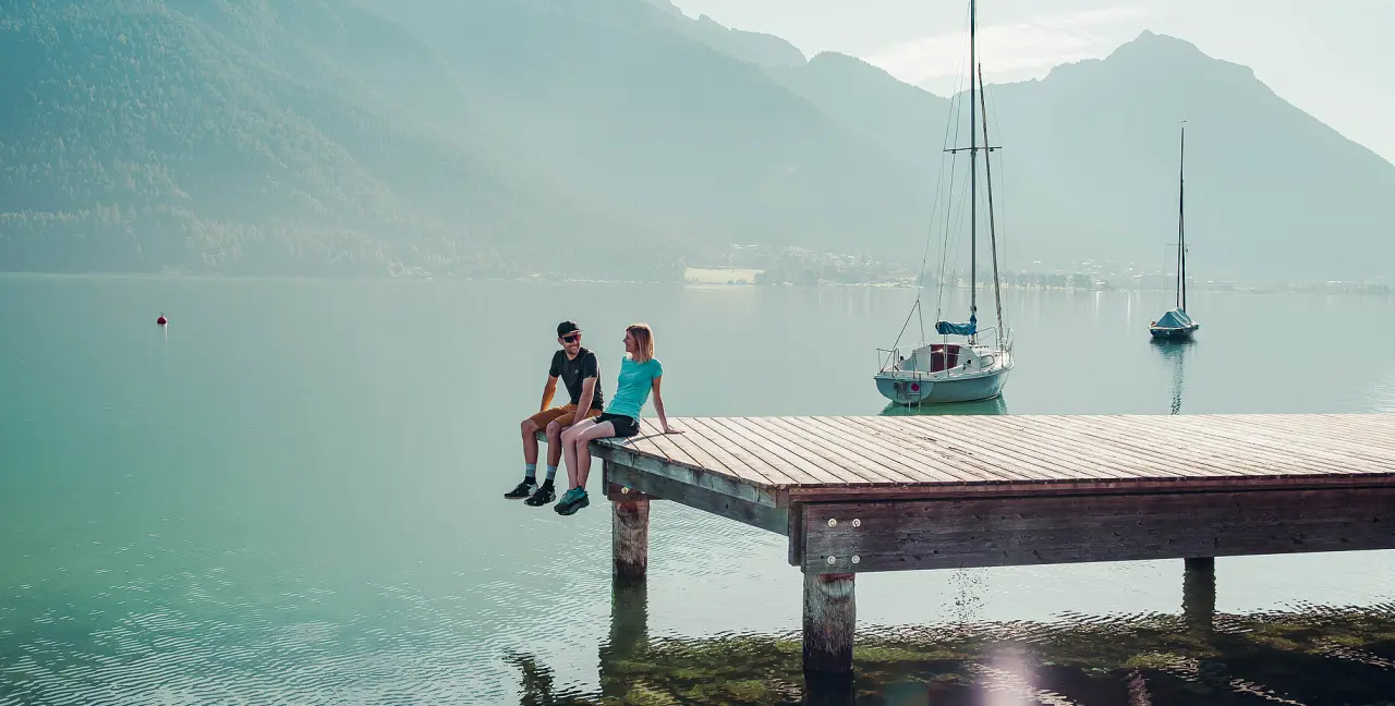 Paar sitzt auf einem Holzsteg am klaren See mit Segelbooten und Bergpanorama beim Hotel St. Georg zum See.