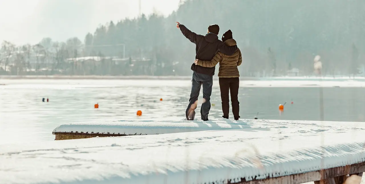 Paar auf verschneitem Steg am See, Blick auf die winterliche Landschaft des Hotel St. Georg zum See.