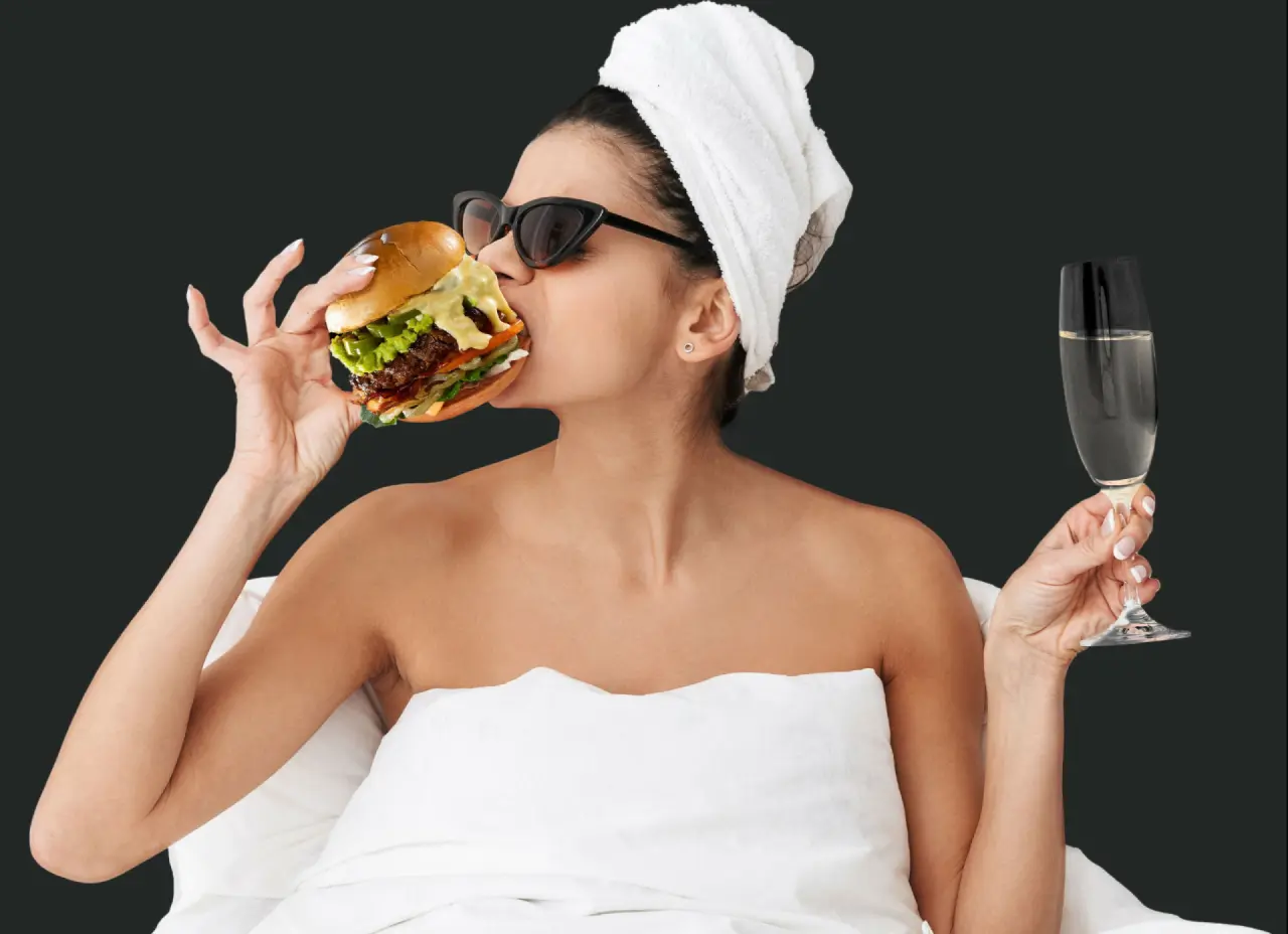 Frau mit Handtuch und Sonnenbrille isst Burger und trinkt Champagner im Hotelzimmer des Loxone Campus.