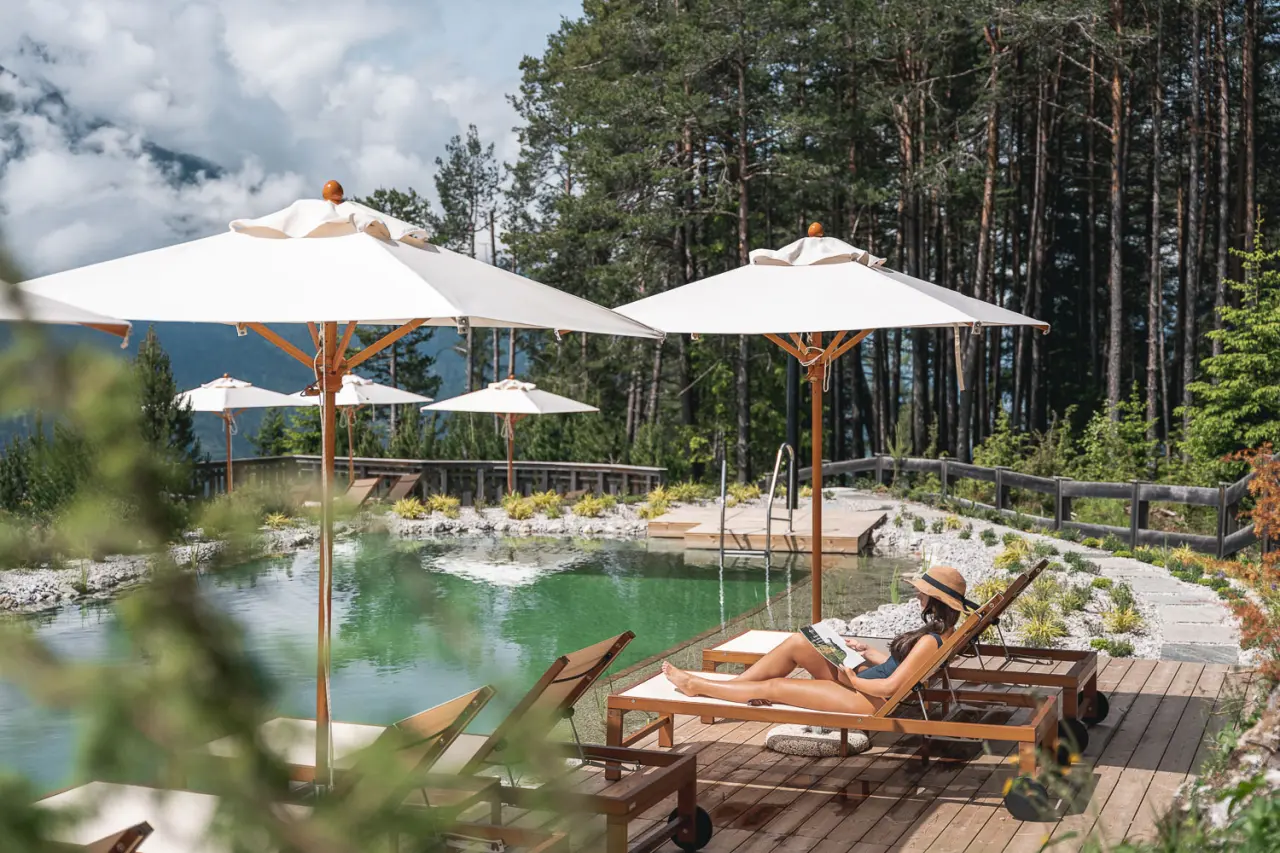 Naturschwimmteich mit Liegestühlen und Sonnenschirmen im Hotel Nidum, umgeben von Wald und Bergen.