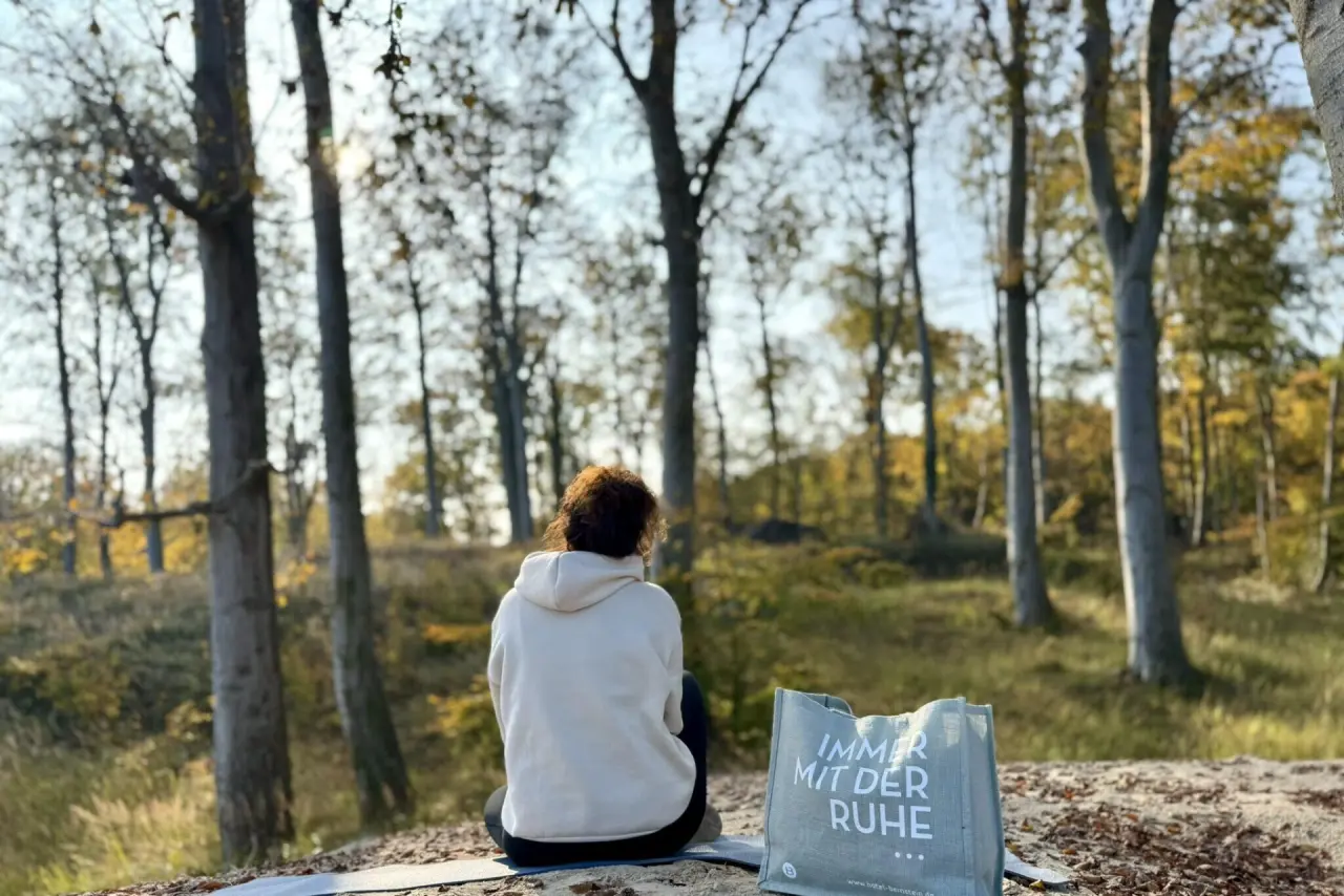 Person meditiert auf einer Matte im herbstlichen Wald, umgeben von Bäumen, mit einer 'Immer mit der Ruhe' Tasche, Hotel Bernstein.