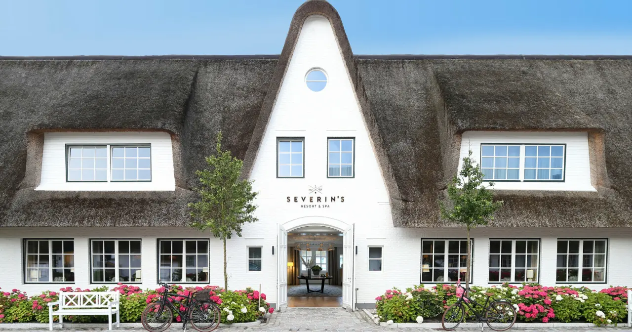 Einladende Aussenansicht des Severin*s Resort & Spa mit Reetdach, weisser Fassade und blühenden Hortensien.