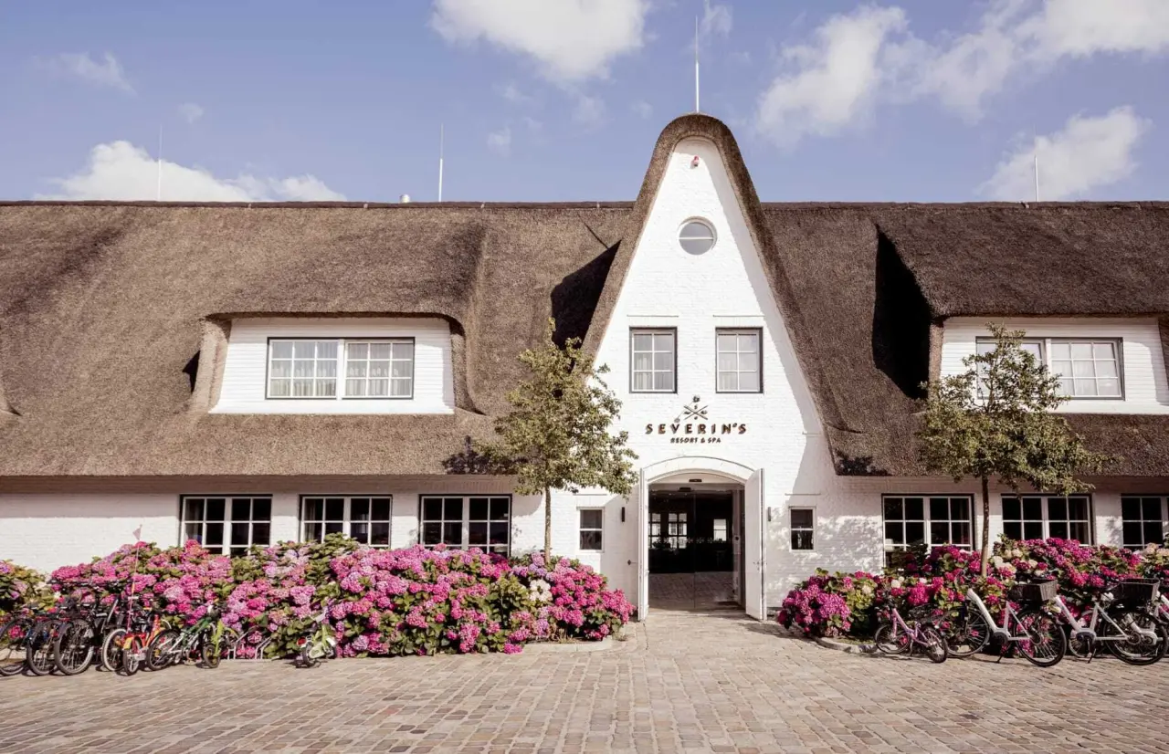 Charmante Aussenansicht des Severin*s Resort & Spa mit Reetdach, pinken Hortensien und Fahrrädern vor dem Eingang.