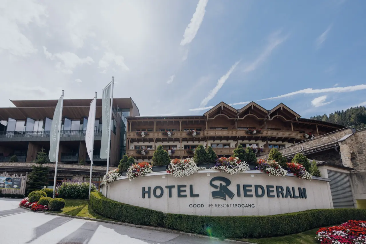 Aussenansicht des Good Life Resort Riederalm in Leogang mit Hotelbeschilderung, traditioneller und moderner Architektur und Blumen.