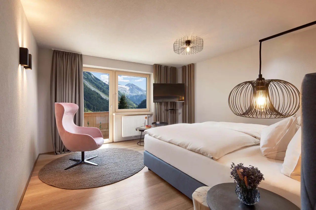 Modernes Hotelzimmer im ADLER INN Tyrol Mountain Resort mit gemütlichem Bett, Sessel und Balkonblick auf die Alpen.