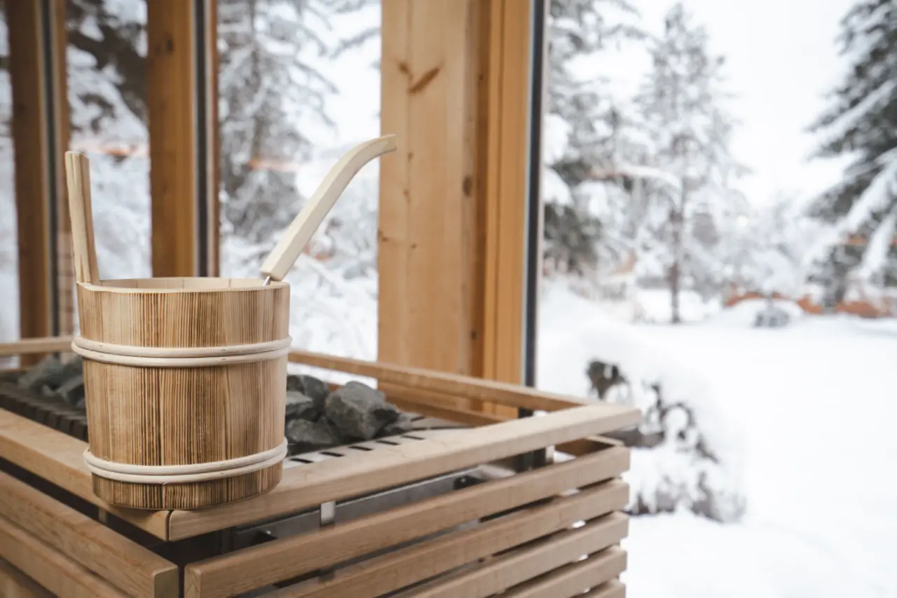 Saunaofen mit Holzeimer und Schöpfkelle im Evident Hotel Prägant. Blick auf verschneite Winterlandschaft.