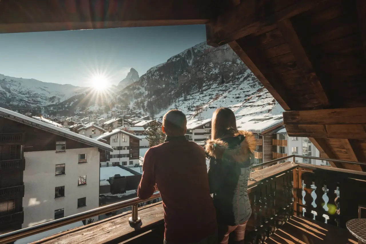 Paar auf Balkon mit Blick auf verschneites Bergdorf und Matterhorn bei Sonnenuntergang im Alpen Resort Hotel.