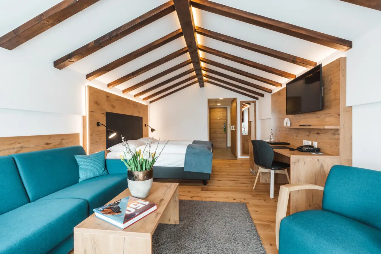 Modernes Hotelzimmer im Alpen Resort Hotel mit Holzbalken, Doppelbett, türkisfarbenem Sofa und Schreibtisch.