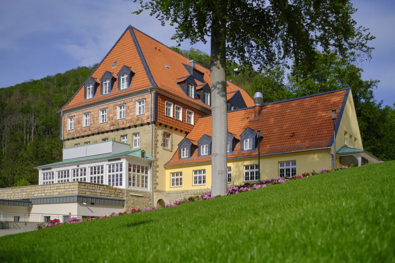 Aussenansicht des sonnenresorts ETTERSHAUS mit rotem Dach, gelben Fassaden und grüner Wiese im Vordergrund, umgeben von Bäumen.