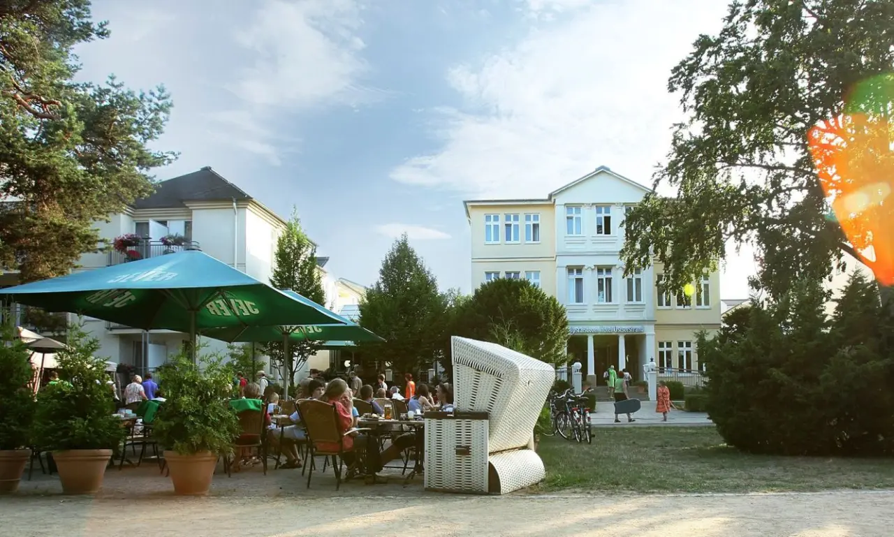 Aussenansicht des Upstalsboom Hotel Ostseestrand mit belebter Terrasse, Strandkorb und grünen Bäumen unter blauem Himmel.