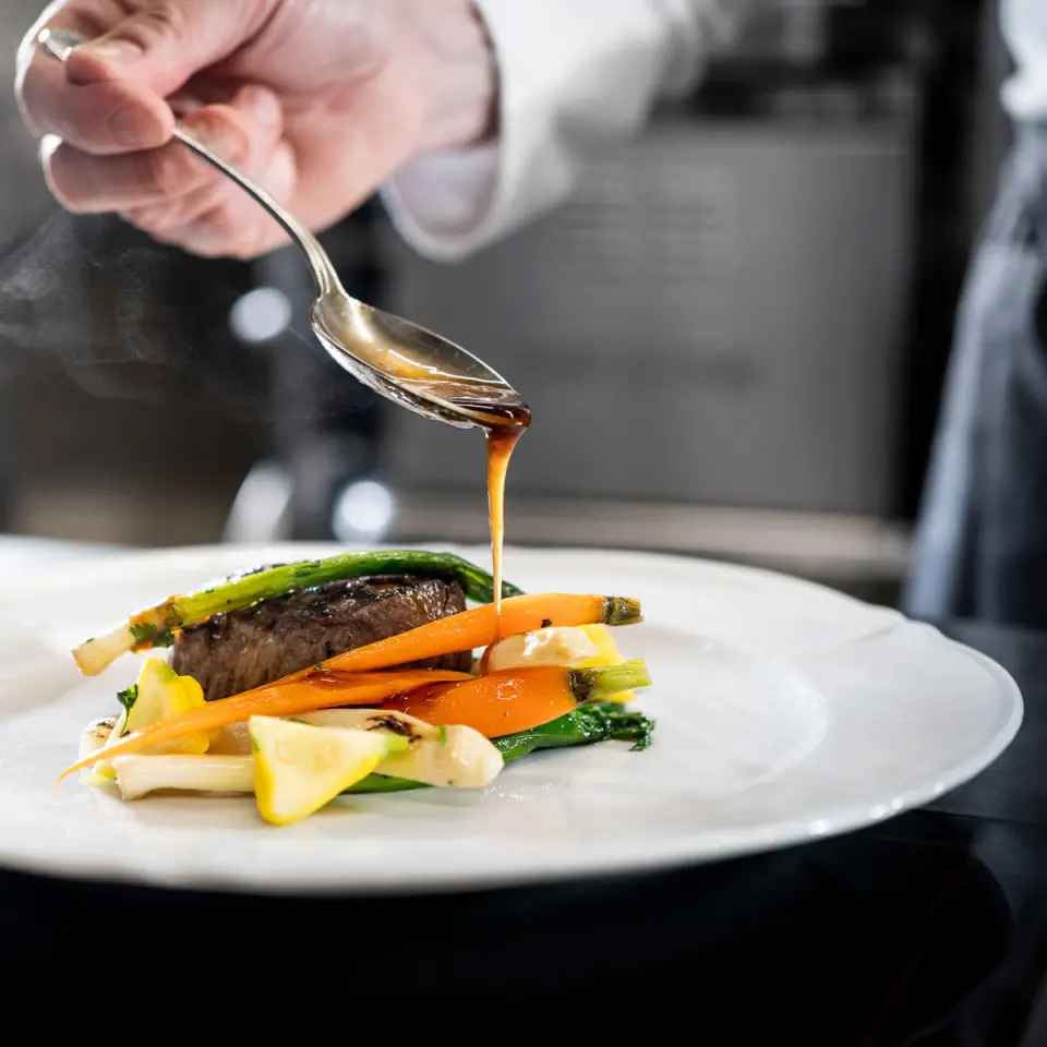 Chef beim Anrichten eines Gourmet-Gerichts mit Fleisch und Gemüse im Restaurant des BLUME. Baden Hotel & Restaurant.