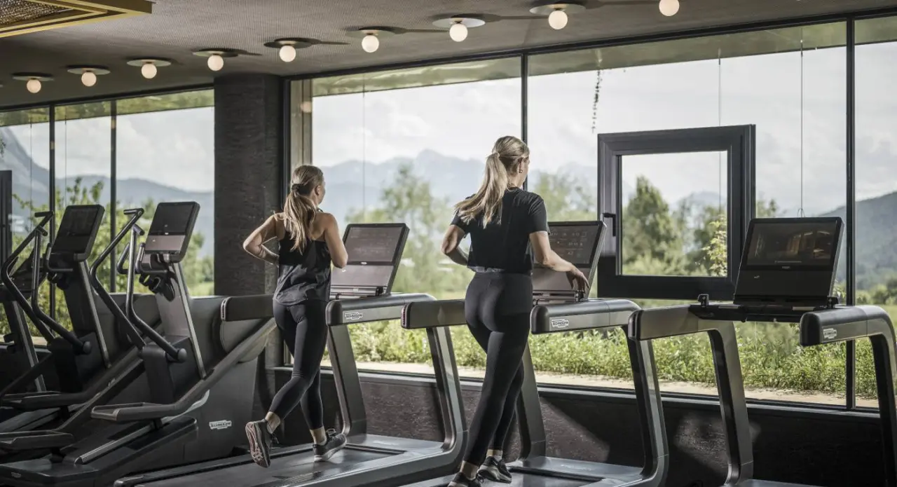 Moderner Fitnessbereich im Alpenresort Schwarz mit zwei Frauen auf Laufbändern und Panoramablick auf die Berge.