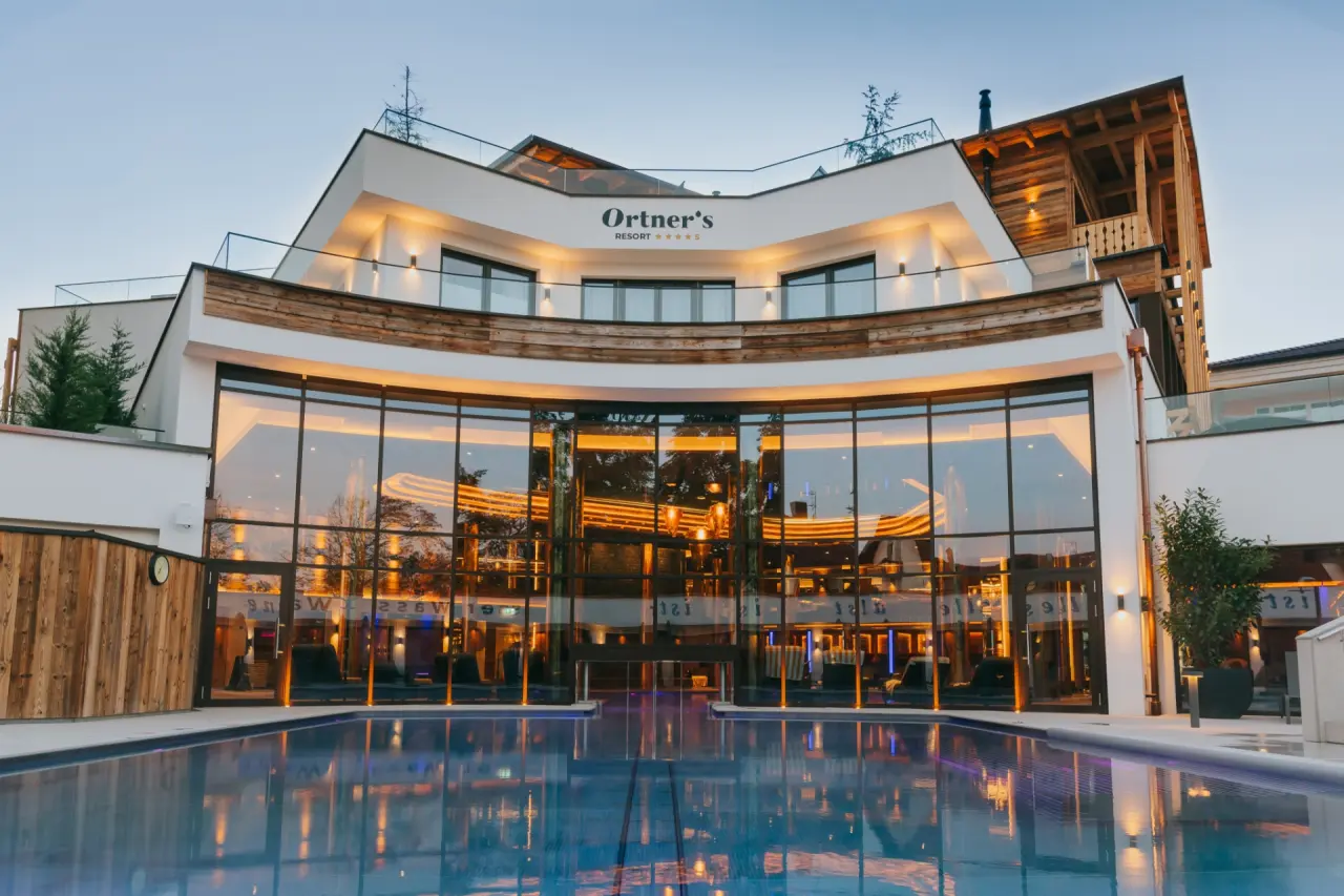 Modernes Ortner's Resort bei Dämmerung mit beleuchteter Glasfassade und Aussenpool. Einladende Wellness-Architektur.