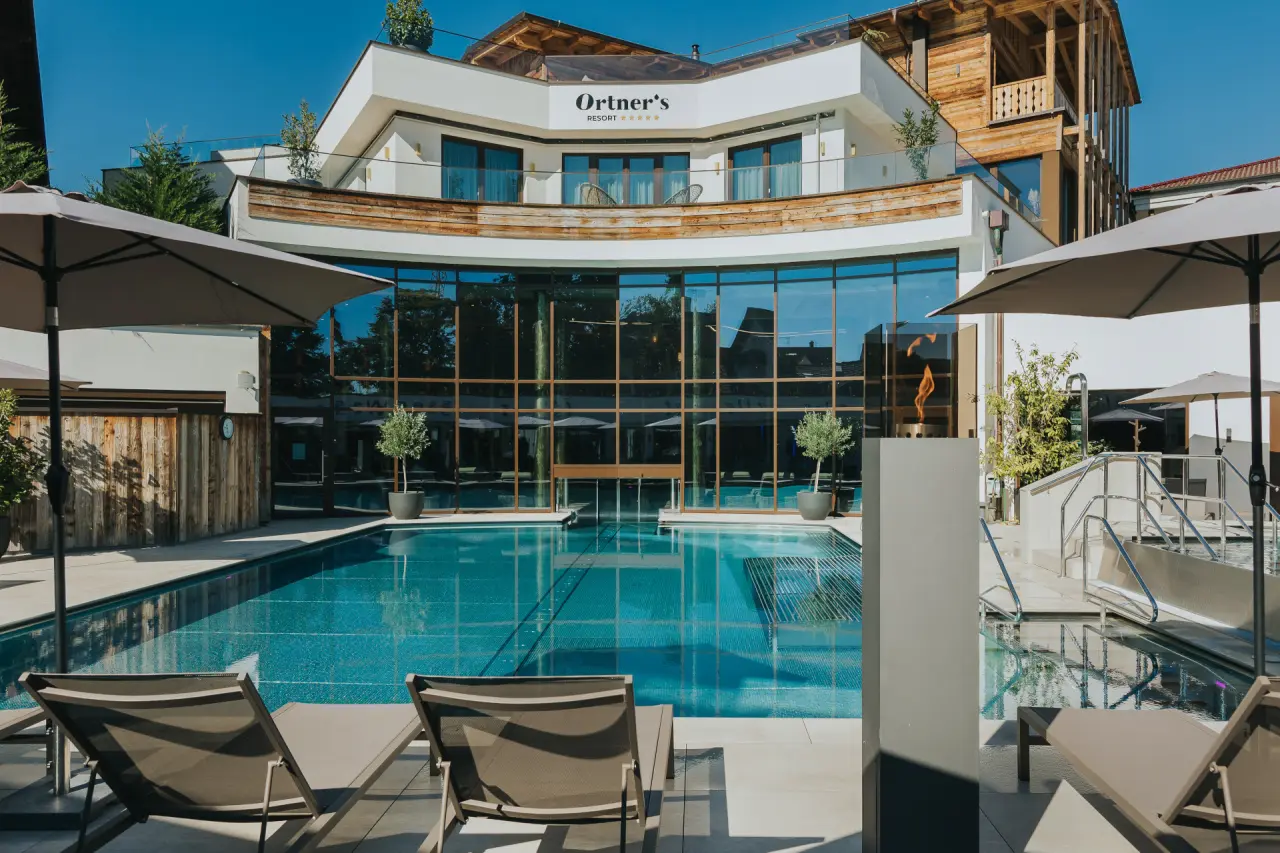 Grosszügiger Aussenpoolbereich mit Sonnenliegen und moderner Hotelarchitektur des Ortner's Resort bei strahlendem Sonnenschein.