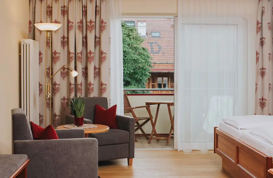 Helles Hotelzimmer im Ortner's Resort mit bequemen Sesseln, Balkon und Blick auf die Dächer und Bäume.