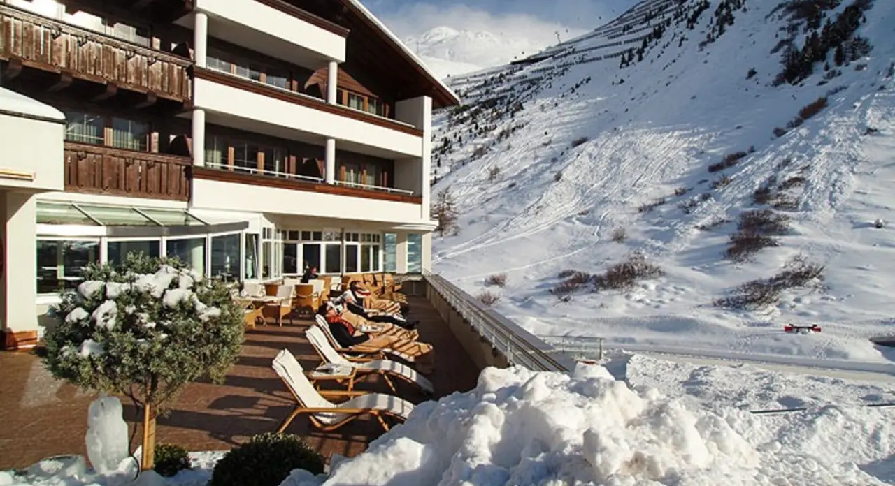 Hotel Alpina Deluxe mit sonniger Terrasse und Liegestühlen im Schnee vor beeindruckendem Bergpanorama.