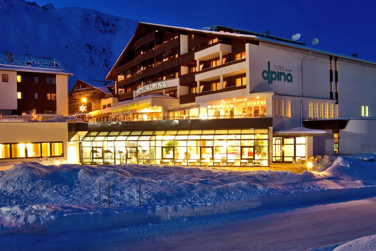 Beleuchtete Aussenansicht des Hotel Alpina Deluxe bei Nacht, eingebettet in eine verschneite Winterlandschaft mit Bergen.