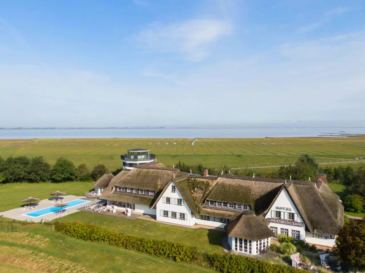 Luftaufnahme des Hotels Lundenbergsand Nordsee Hideaway und Spa mit Reetdach, Aussenpool und Blick auf das Wattenmeer.