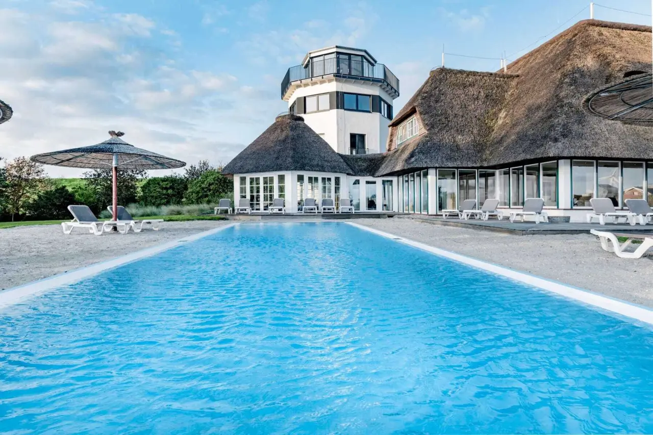 Aussenpool mit Liegen und Reetdach-Hotelgebäude im Lundenbergsand Nordsee Hideaway und Spa.