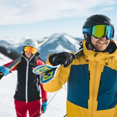 Zwei lächelnde Skifahrer mit Ausrüstung vor verschneiter Bergkulisse am Sporthotel Ellmau.