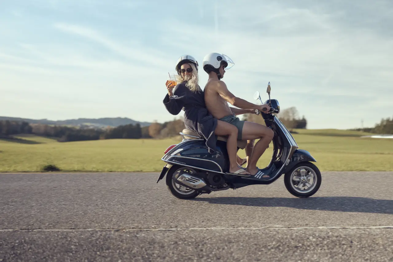 Paar im Bademantel auf Vespa Roller mit Drink vor ländlicher Kulisse, Hotel Guglwald.