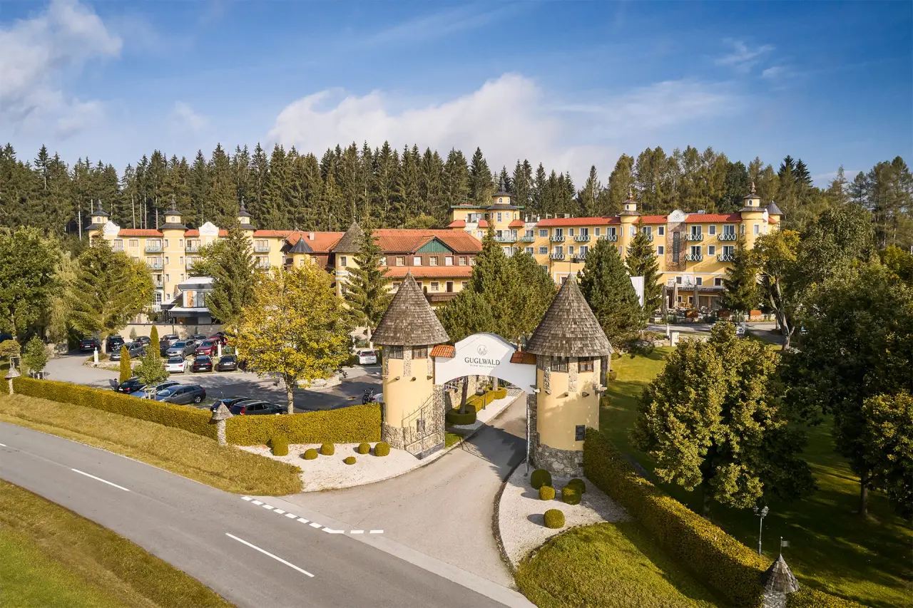 Panoramablick auf das Hotel Guglwald, ein Wellnesshotel mit markantem Eingangstor und gelber Fassade, umgeben von Wald.