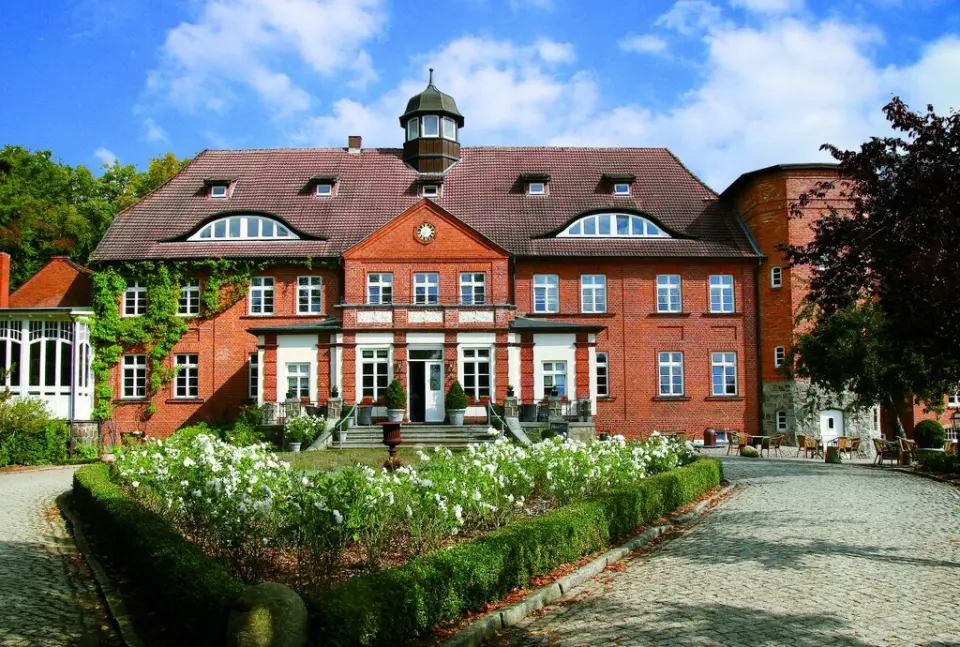 Majestätisches Schloss Basthorst mit roter Backsteinfassade, grünen Ranken und blühendem Vorgarten unter blauem Himmel.