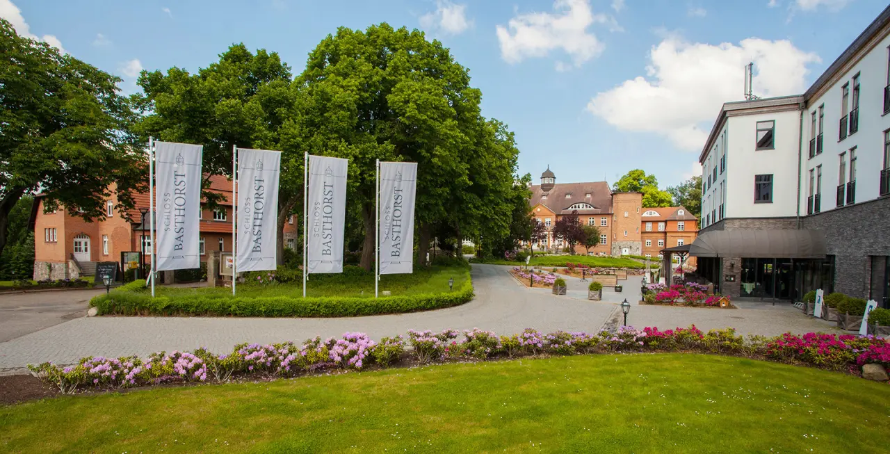 Aussenansicht des Schlosshotels Schloss Basthorst mit gepflegter Auffahrt, gruenen Baeumen und bunten Blumenbeeten.