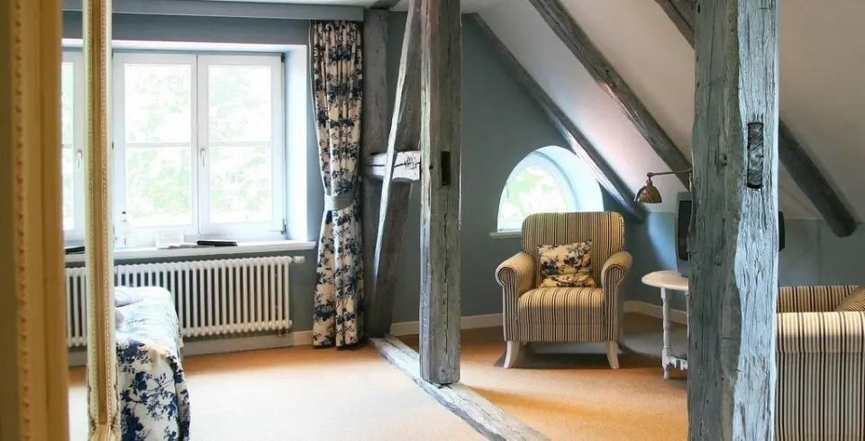 Gemütliches Dachzimmer im Schloss Basthorst mit Holzbalken, gestreiftem Sessel und Fensterblick ins Grüne.
