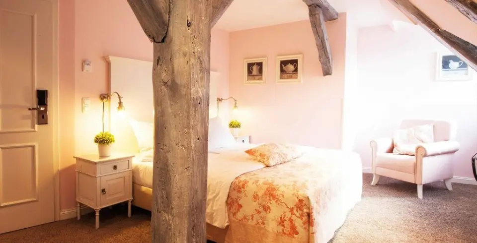 Gemütliches Hotelzimmer im Schloss Basthorst mit Doppelbett, rosa Wänden, Holzbalken und Sessel.
