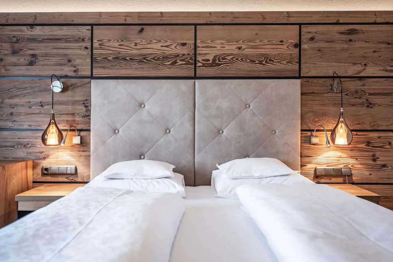 Gemütliches Doppelbett mit Holzwand und Leselampen im modernen Zimmer des Naturhotel Alpenblick.