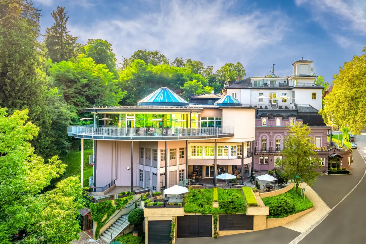 Aussenansicht des Hotel Allmer mit moderner Architektur, grünen Terrassen und umliegendem Wald unter blauem Himmel.