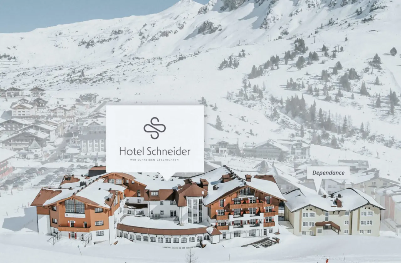 Winterpanorama des Hotel Schneider mit verschneiten Gebäuden und umliegendem Dorf vor einer beeindruckenden Bergkulisse.