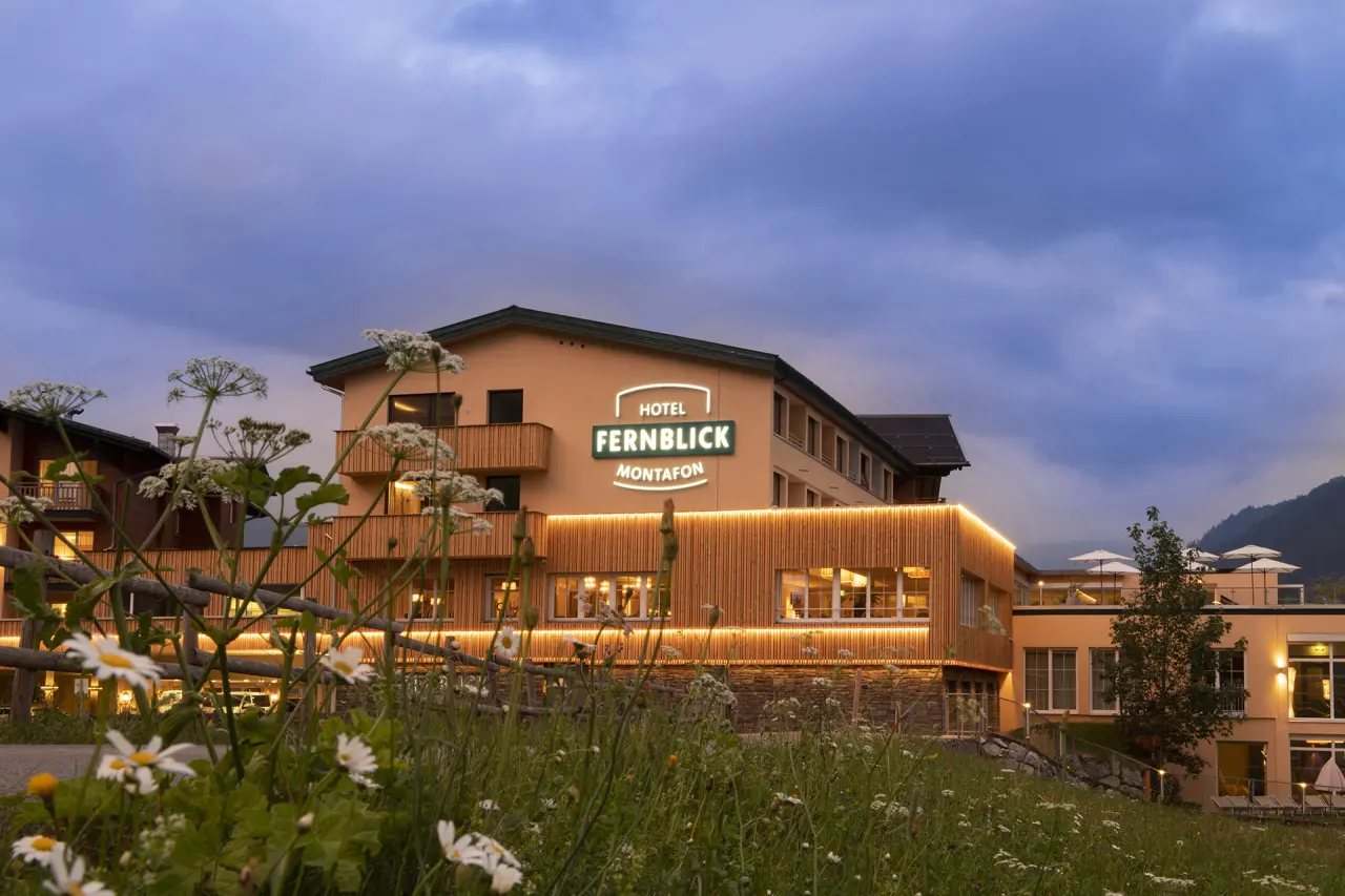 Abendansicht des Hotel Fernblick Montafon mit beleuchteter Fassade und blühender Wiese im Vordergrund.