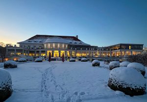 ATLANTIC Grand Hotel Travemünde bei Dämmerung im Winter. Das elegant beleuchtete Hotelgebäude mit verschneitem Garten.